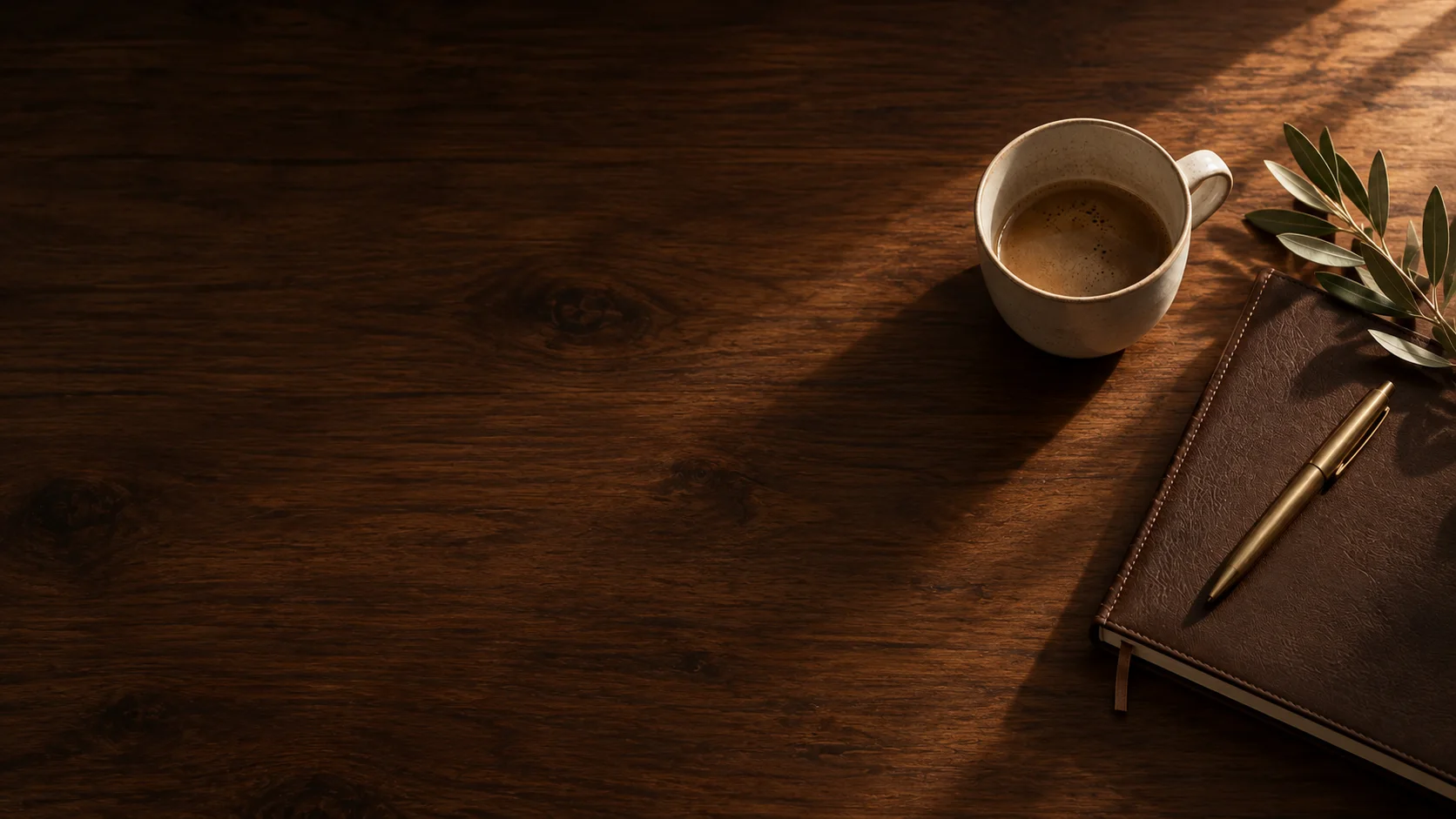 Mesa de madera con una taza de café, un cuaderno de piel, una pluma de latón y una rama de olivo bajo la luz de la mañana